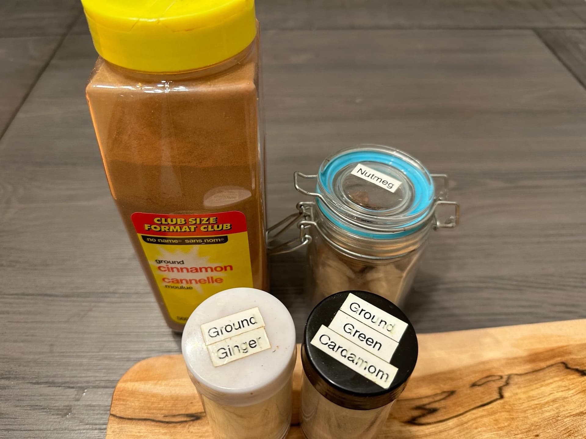 Apple pie spice ingredients including ground cinnamon jar, ground ginger container, green cardamom jar, and cinnamon bottle on wooden surface.