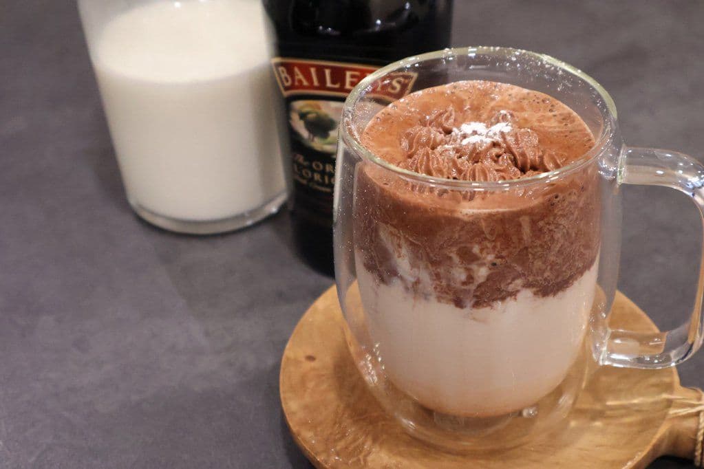 Whipped Baileys hot chocolate in a clear mug with cocoa powder dusting, beside cream and Baileys bottle.