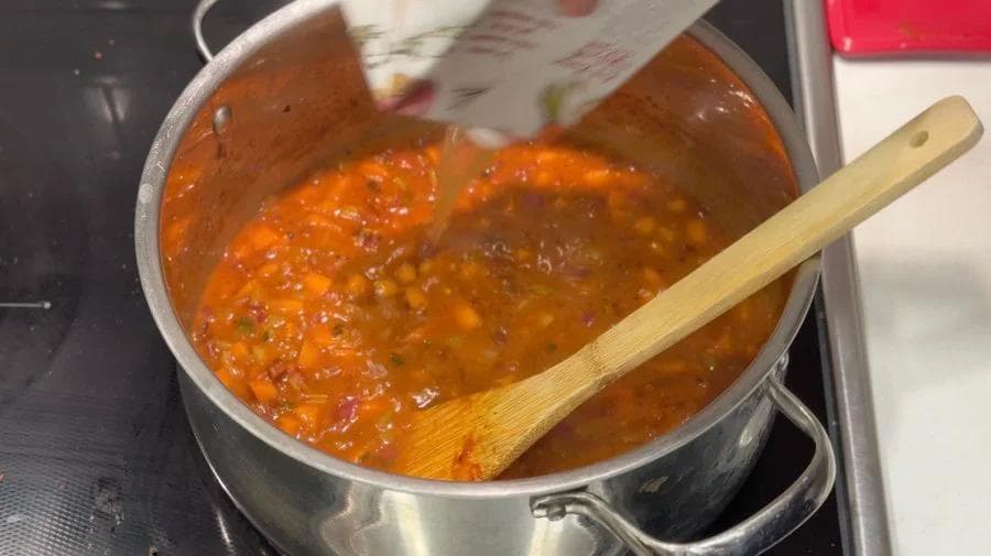 Tomato paste and broth simmering in a pot with a wooden spoon, creating an orange-red harissa stew base.