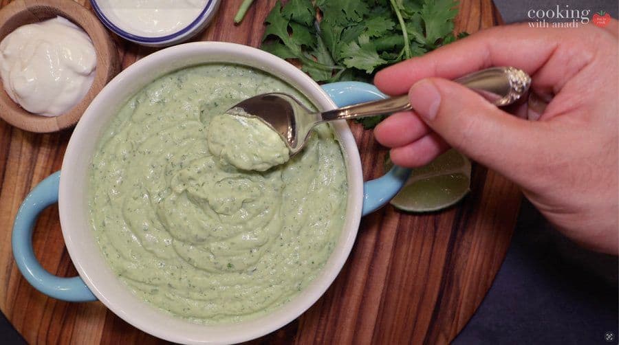 Creamy avocado lime crema being spooned from a white bowl with blue handles, showing its smooth, pale green texture.
