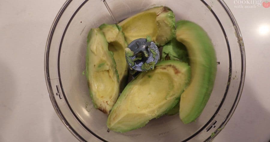 Halved avocados with pit in a food processor bowl, ready to blend into crema sauce.