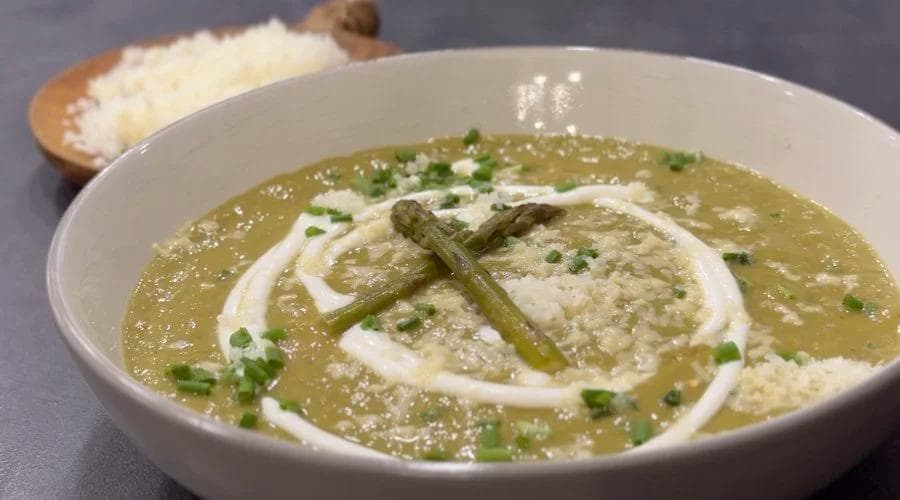 Creamy asparagus soup garnished with sour cream swirl, asparagus tips, chives, and cheese in a white bowl.