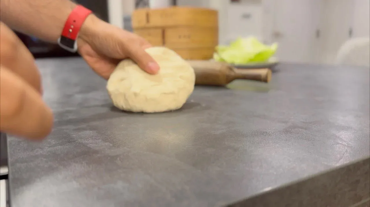 Hand shaping a round momo dough disk on a dark work surface during preparation.