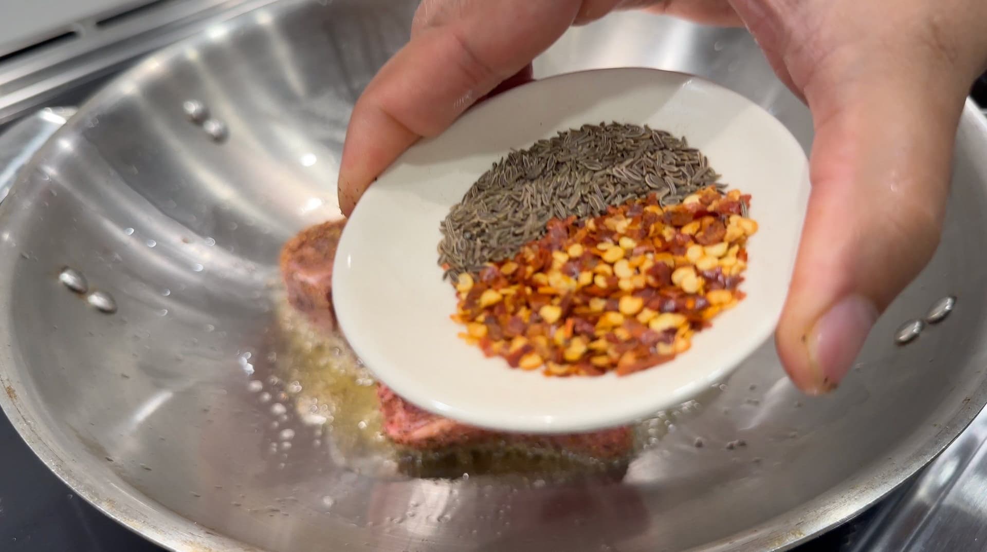 Hand holding white plate with cumin seeds, red chili flakes, and diced vegetables over a metal wok.