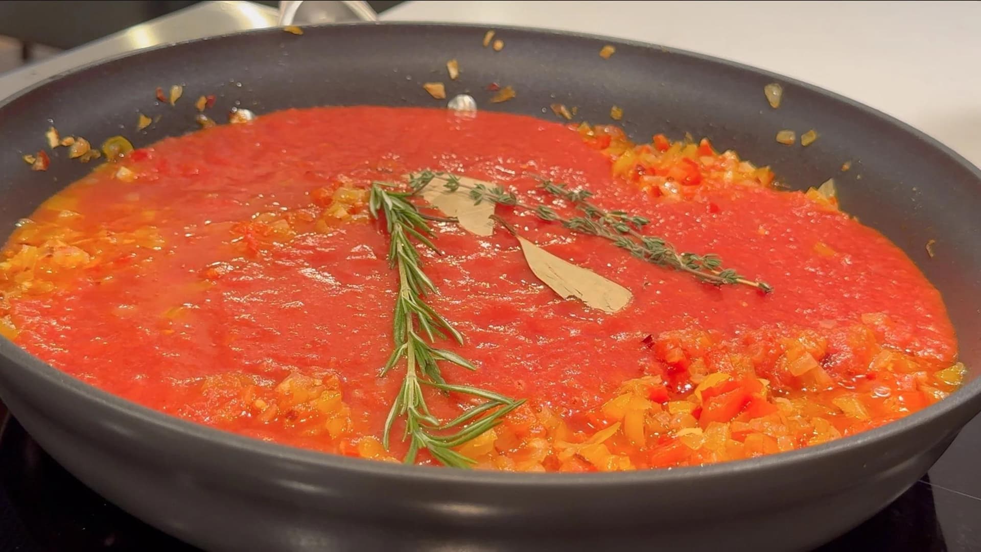 Tomato piperade simmering in a skillet with fresh rosemary, thyme sprigs, and a bay leaf.