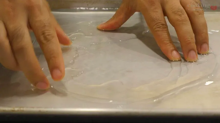 Hands softening a rice paper wrapper in water on a white plate for dumpling preparation.