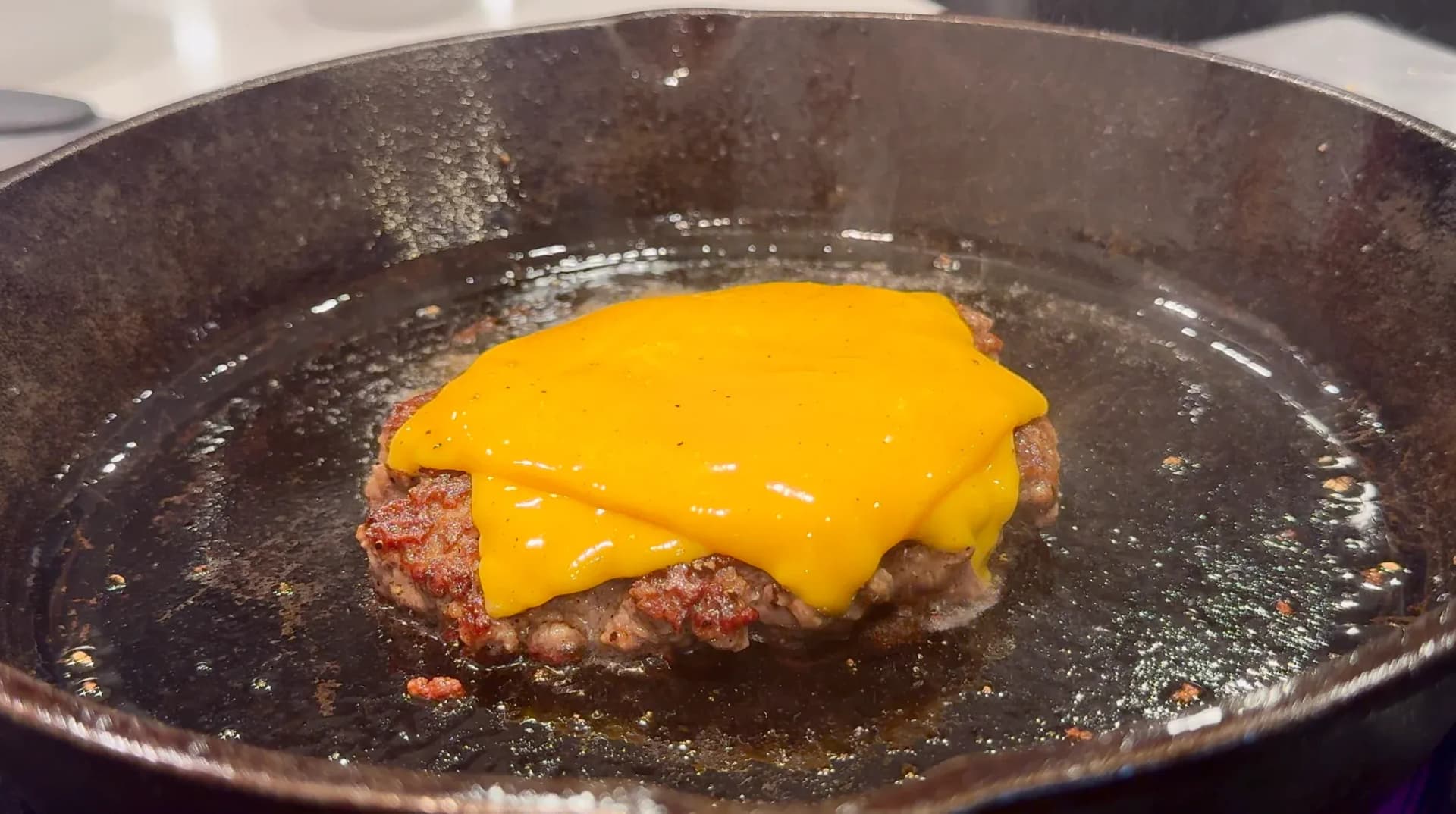 Burger patty with melted yellow cheese cooking in a dark skillet with oil.