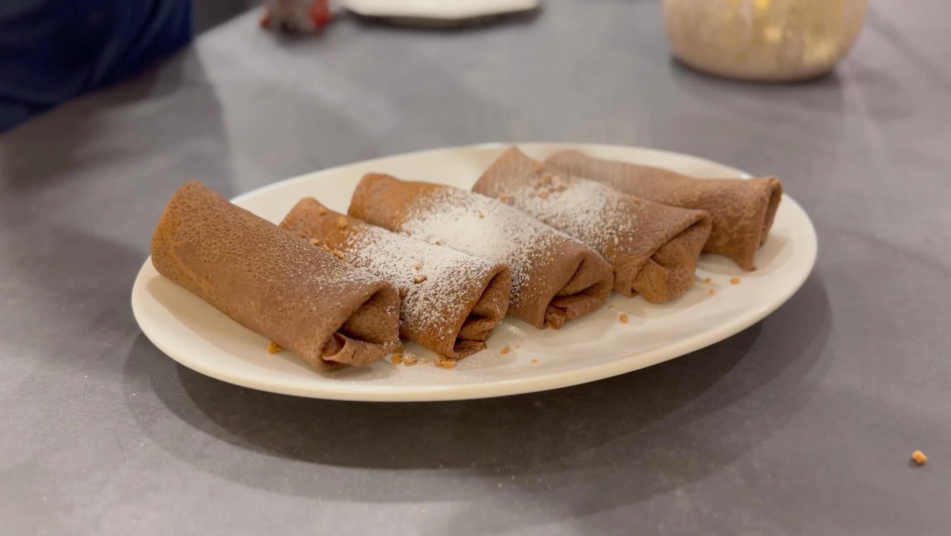 Gluten-free chocolate crepes rolled and dusted with powdered sugar on a white plate.