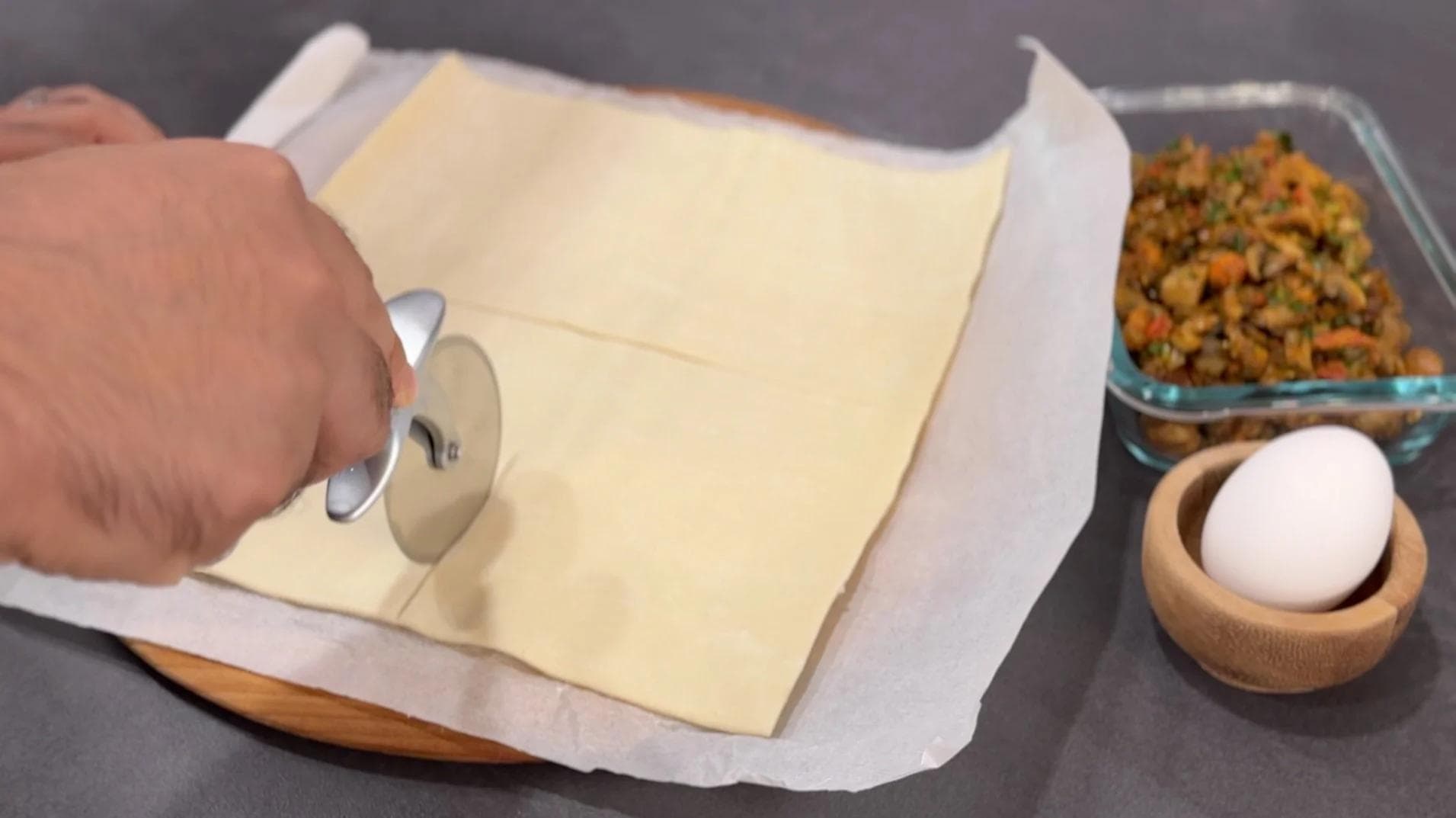 Unrolled puff pastry being cut into squares with a pizza wheel, with egg and mushroom filling nearby.