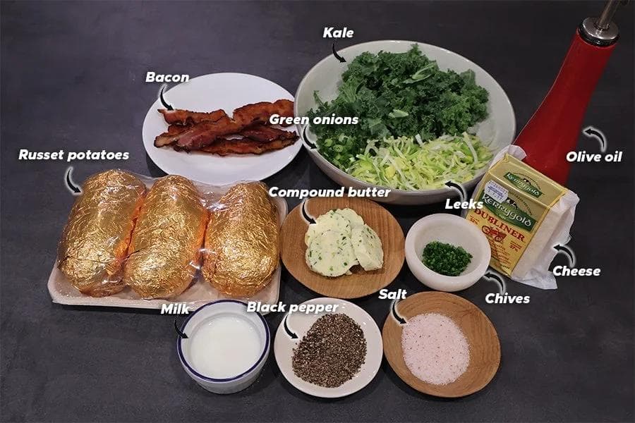 Ingredients for Irish style twice baked potatoes arranged on dark surface including russet potatoes, bacon, kale, leeks, cheese, and seasonings.