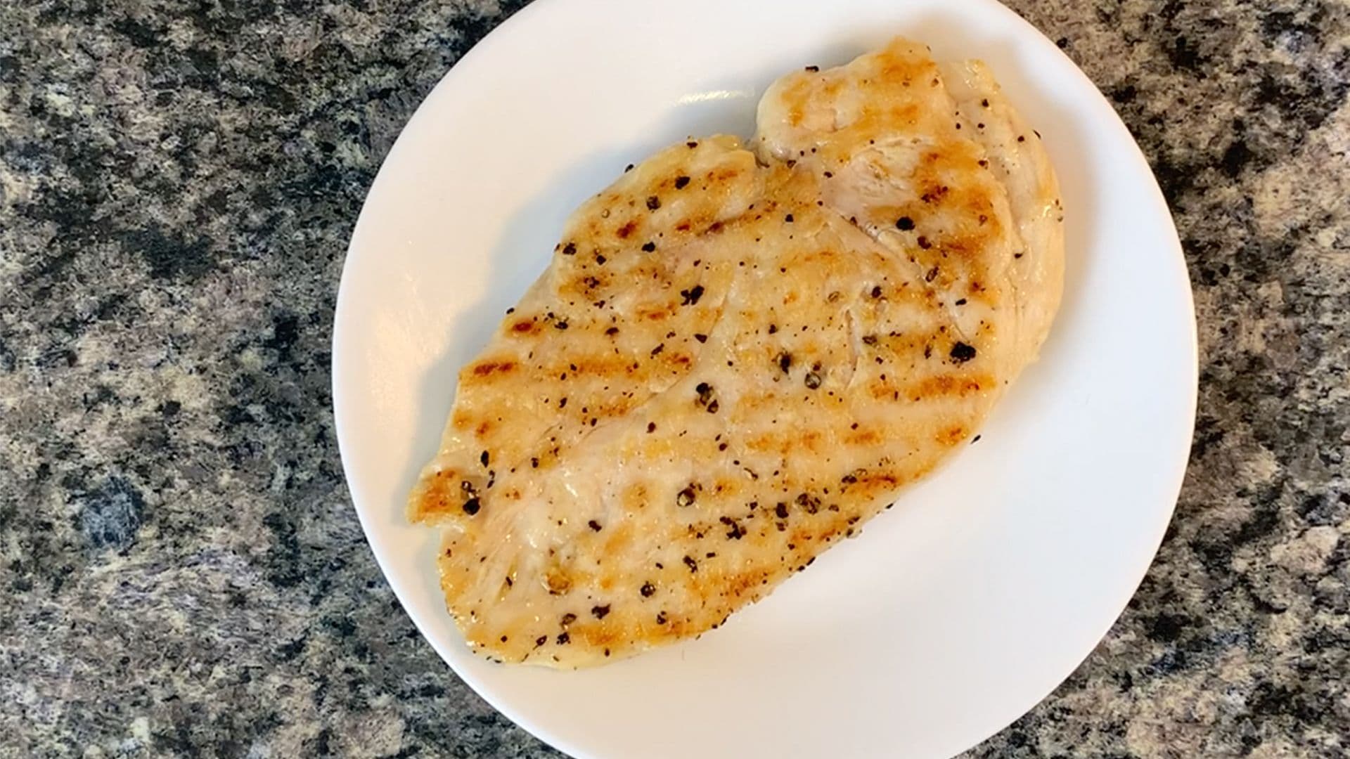 Grilled chicken breast on white plate with black pepper seasoning and golden-brown sear marks.