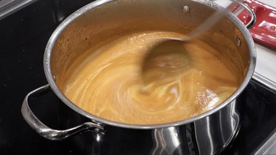 Smooth carrot and ginger soup with coconut milk being stirred in a stainless steel pot on the stovetop.