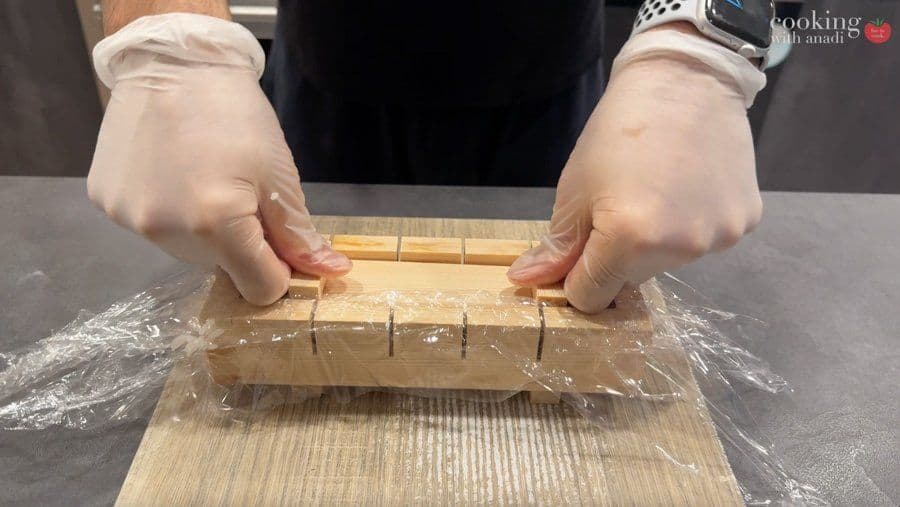 Hands pressing down on wooden mold with plastic-wrapped sushi using a pressing insert to compact it firmly.