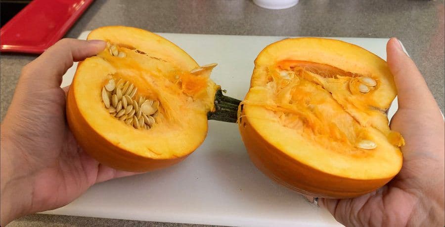 Hands holding two halves of a halved pumpkin with seeds and fibers removed, showing the hollowed orange flesh.