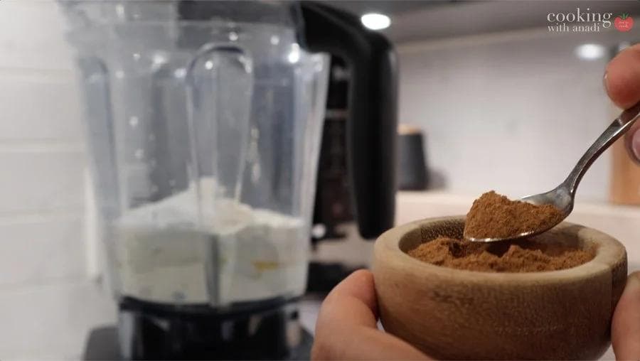 Measuring ground cinnamon with a spoon into a wooden bowl next to a blender with crepe batter.