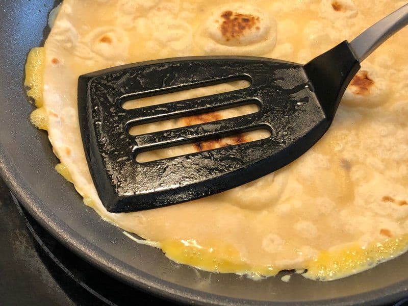 Black spatula pressing cooked eggs onto paratha in a skillet during egg paratha preparation.
