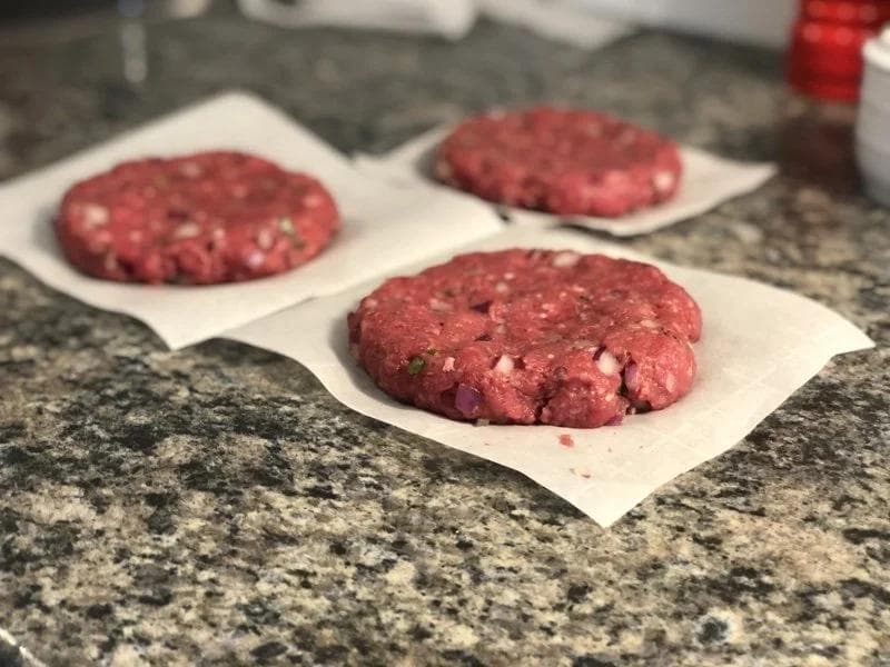 Three raw beef burger patties on parchment paper ready for grilling on a granite countertop.