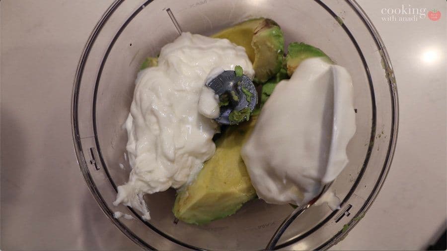 Food processor containing avocado halves, sour cream, and lime with dark seeds visible during blending.