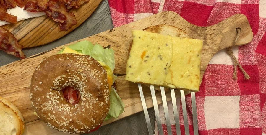 Assembled breakfast bagel sandwich with scrambled egg, bacon, and lettuce on toasted sesame seed bagel on wooden board.