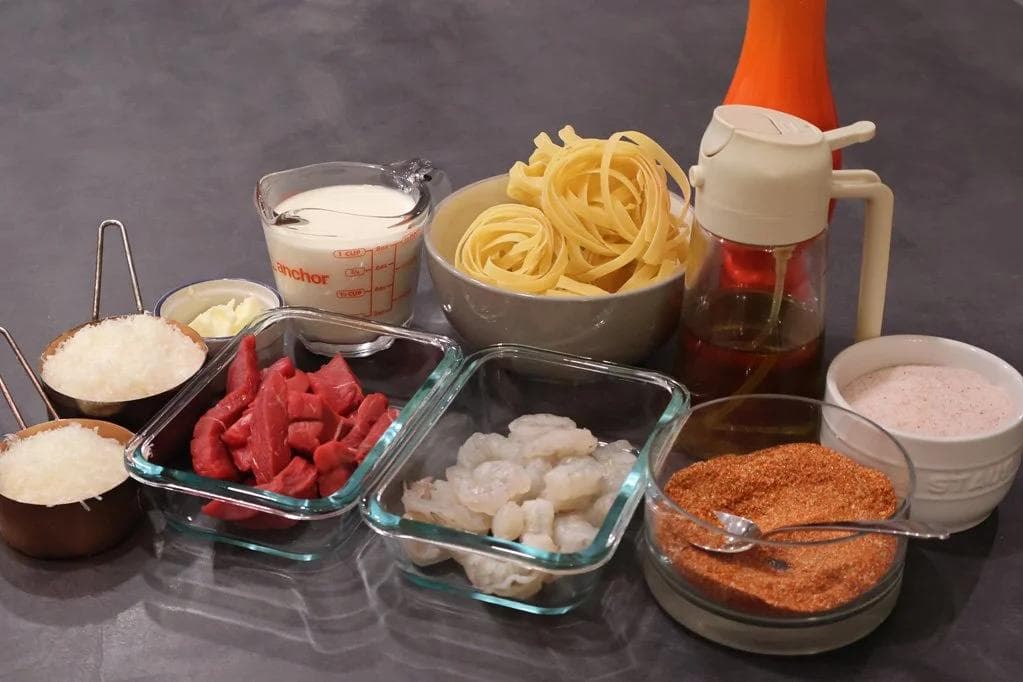Ingredients for Cajun Steak and Shrimp Alfredo arranged on gray surface including pasta, cream, spices, shrimp, and seasonings.