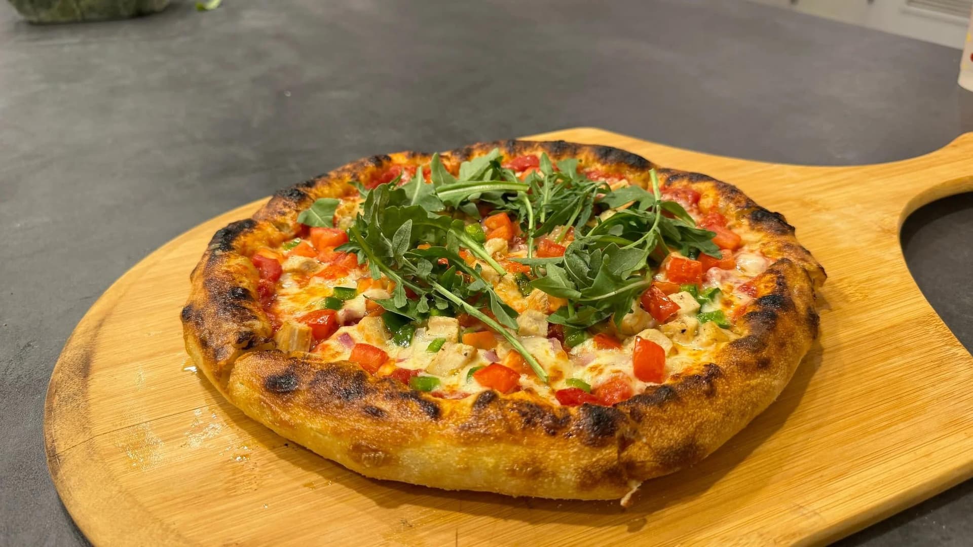Wood-fired pizza with charred crust topped with fresh arugula, red peppers, and melted cheese on wooden serving board.