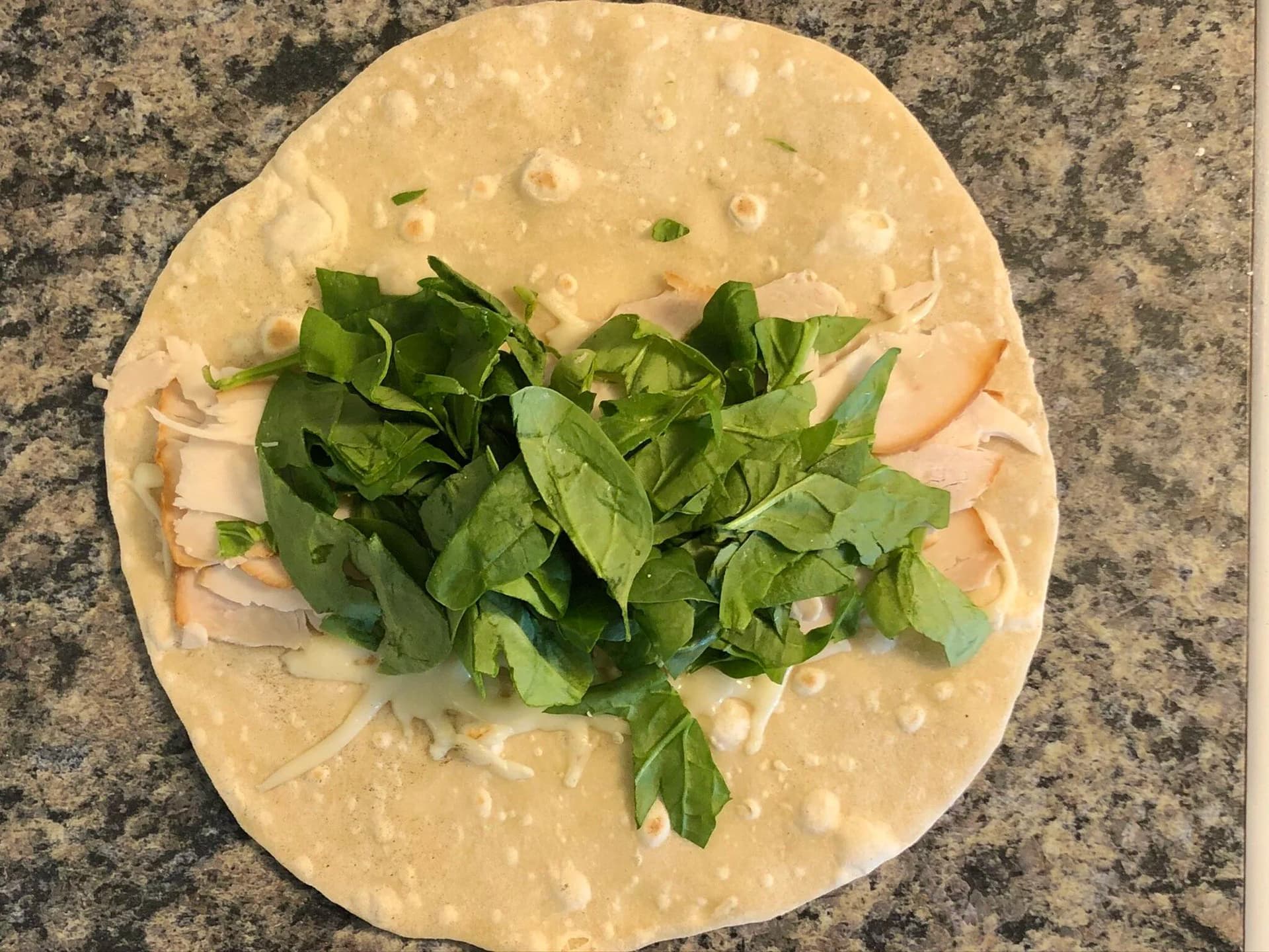 Tortilla wrap topped with melted cheese, sliced deli turkey, and fresh spinach on a granite countertop.