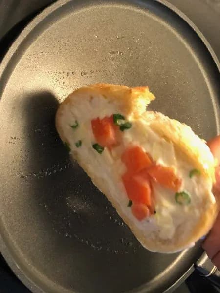 Hand holding a buttery pan-fried cheese toast with melted cheese, diced red peppers, and green onions over a non-stick pan.