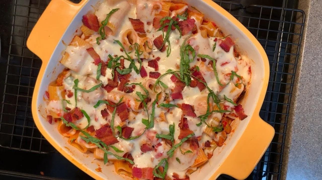 Baked rigatoni pasta with melted cheese, bacon bits, and fresh basil in an orange baking dish.