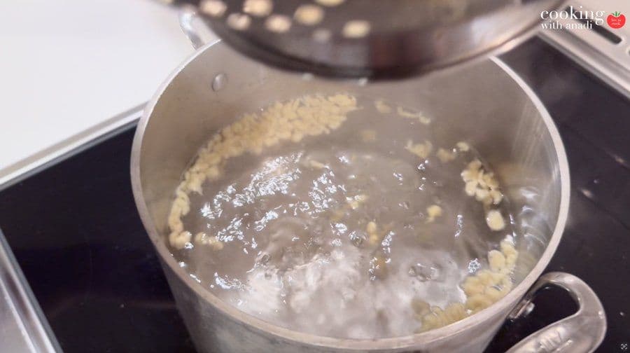 Spätzle batter being pressed through a colander into boiling water, with some pieces already floating.