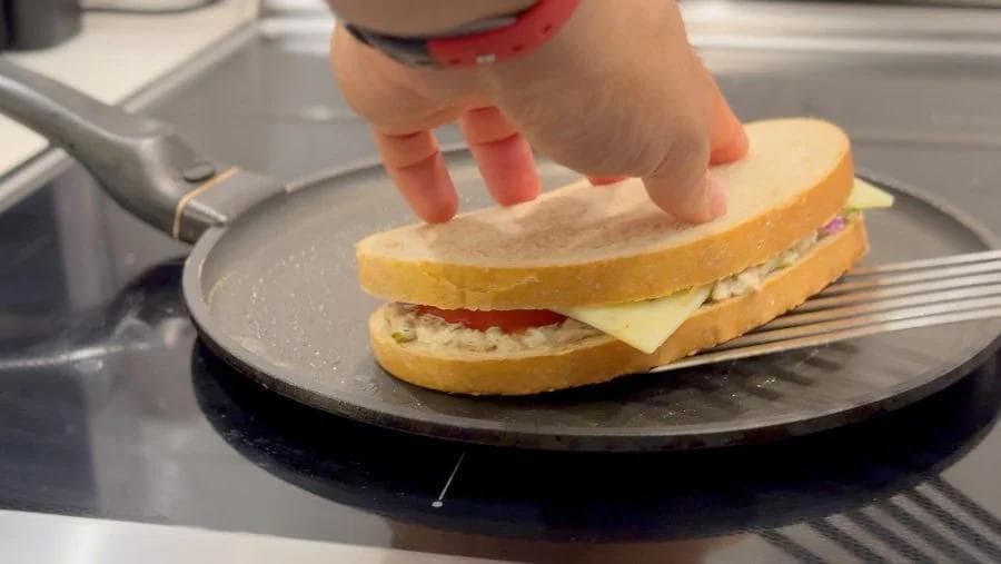 Hand flipping a golden-brown tuna melt sandwich with melted cheese on a non-stick pan.