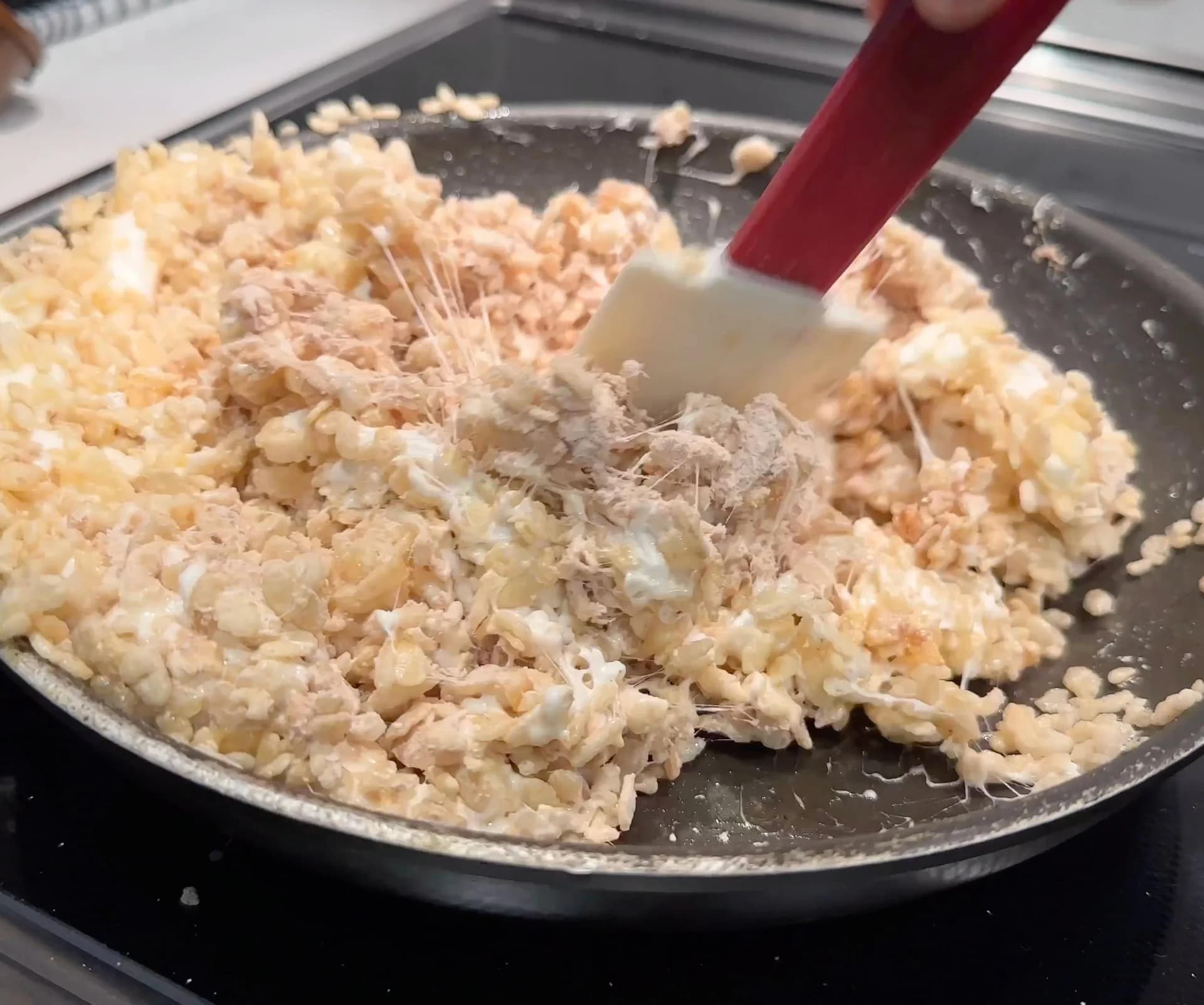 Protein powder and rice cereal being stirred together in a dark skillet with a red spatula.