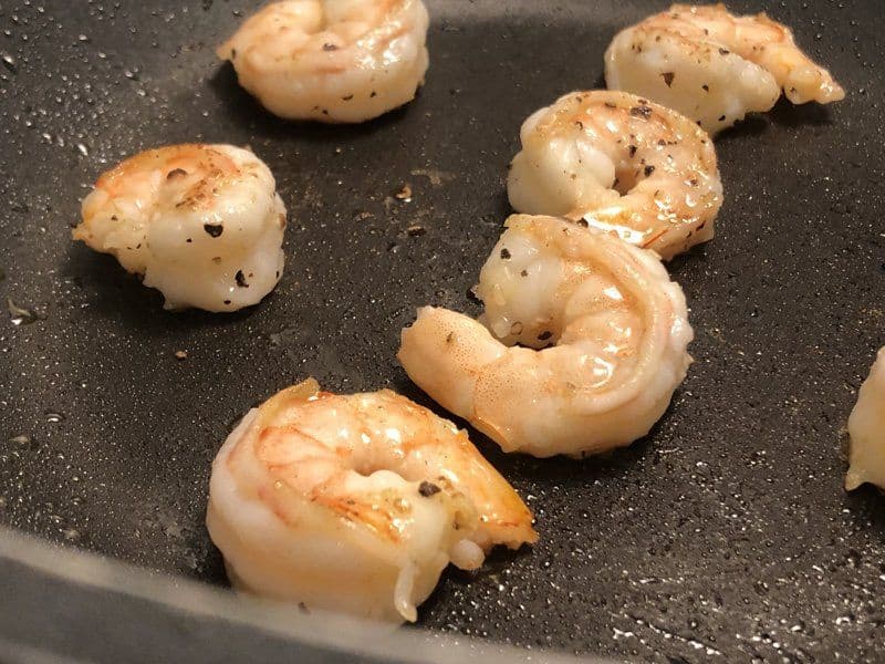 Seared shrimp with black pepper and char marks cooking in a dark pan with oil.