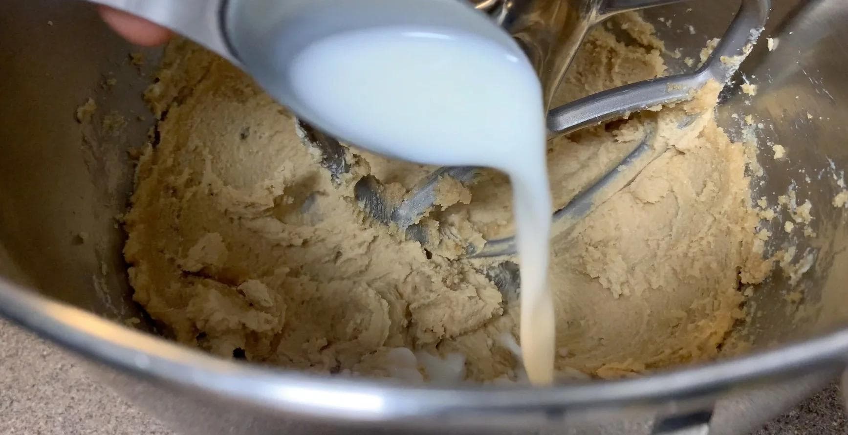 Milk being poured into tan-colored cookie dough in a mixing bowl with a paddle attachment.