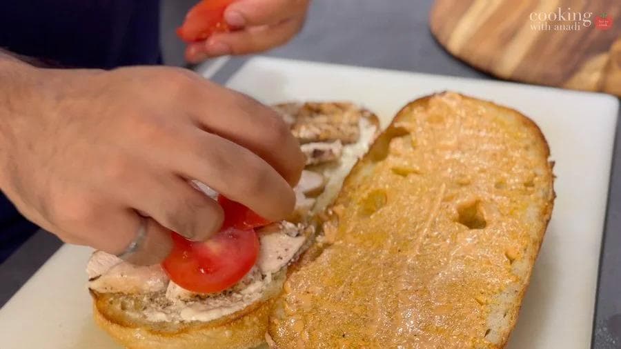 Hand placing a tomato slice on grilled chicken and dressing spread on toasted bread for a Caesar sandwich.