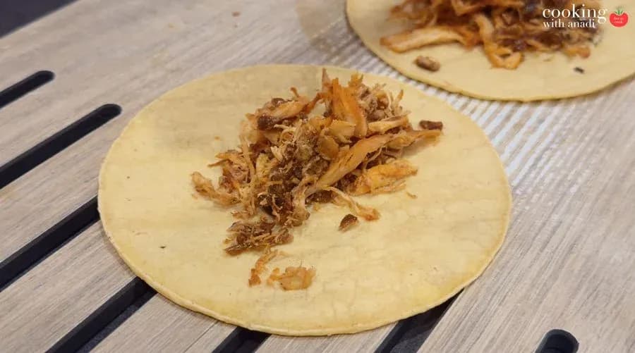 Two corn tortillas on a wooden surface, each topped with seasoned shredded chicken, ready to be rolled into taquitos.