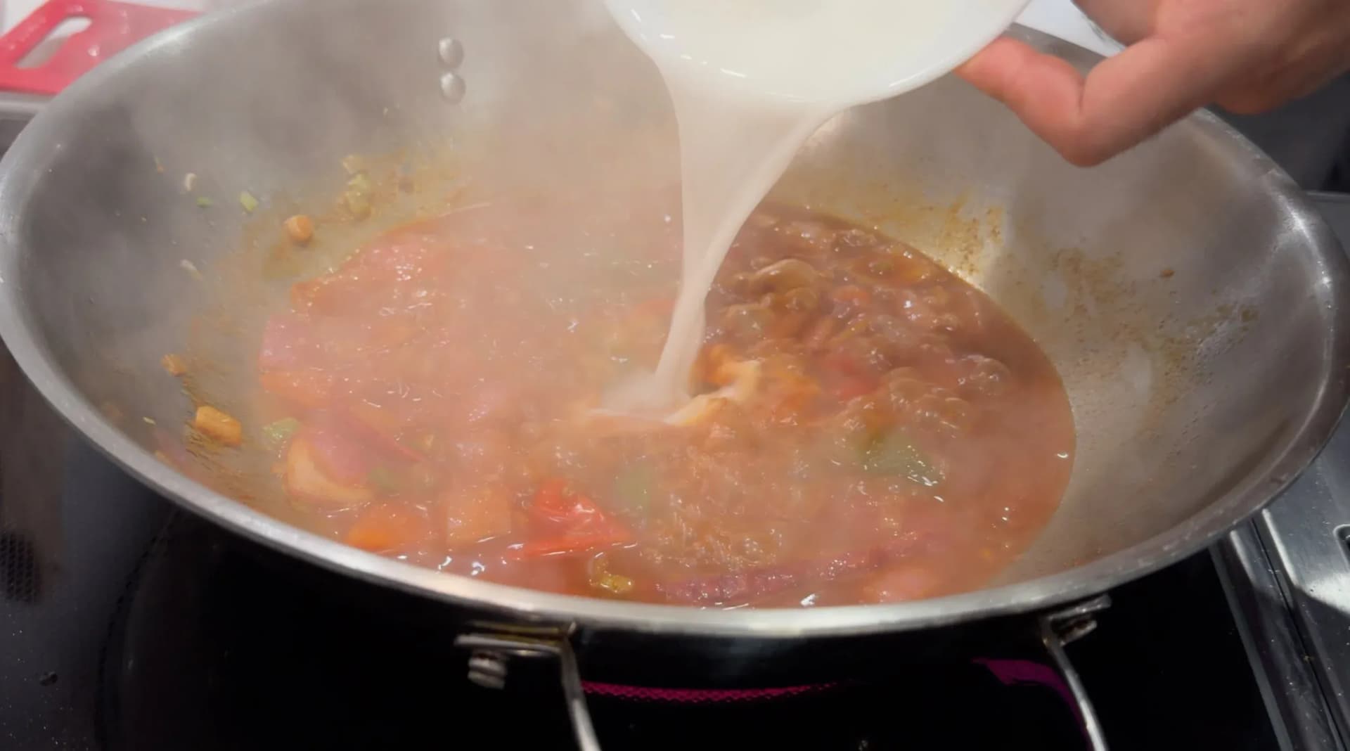 Cornstarch slurry being poured into a simmering tomato-based sauce in a stainless steel wok.