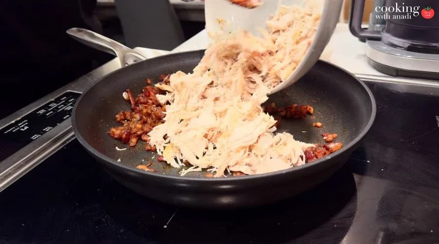 Shredded chicken being poured from a bowl into a skillet with sautéed garlic, onions, and paprika.