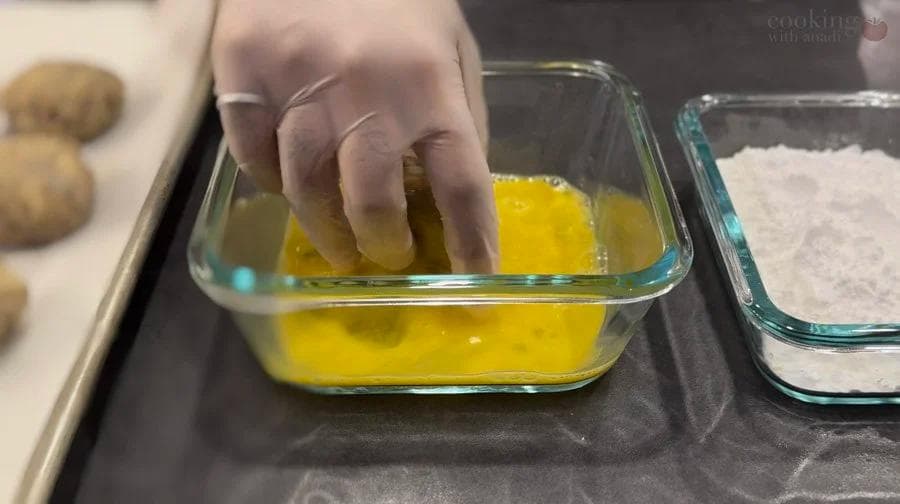 Hand dipping potato fritter into beaten egg wash in a glass bowl before frying.