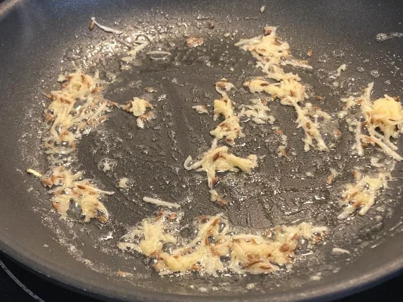 Cumin seeds and ginger-garlic paste cooking in butter in a dark pan, aromatic and beginning to brown.