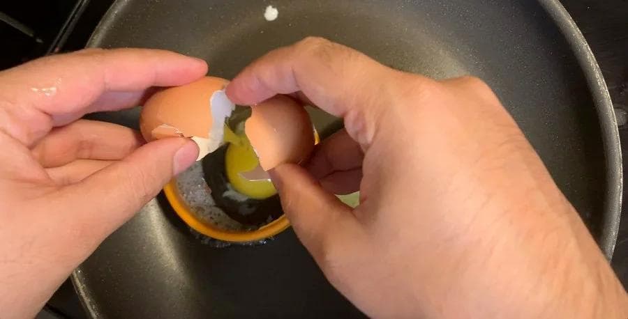 Cracking an egg into a metal ring mold on a buttered pan to shape the egg into a round disk.