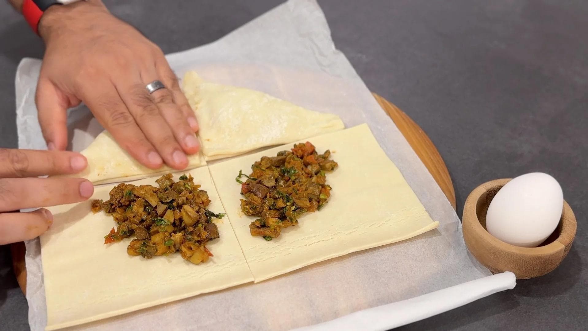 Folding puff pastry squares diagonally over mushroom filling to form triangular pastries, with egg and wooden bowl nearby.