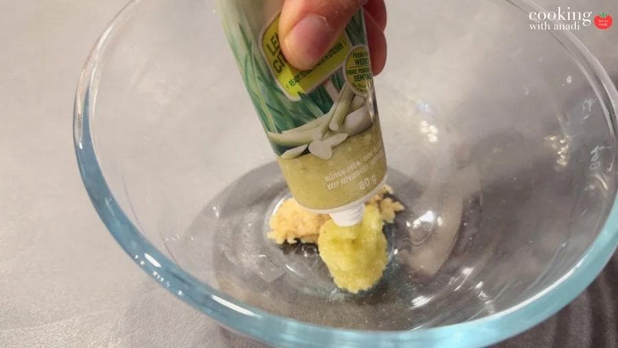 Squeezing lemongrass paste from a tube into a glass bowl with crushed garlic and white pepper paste.