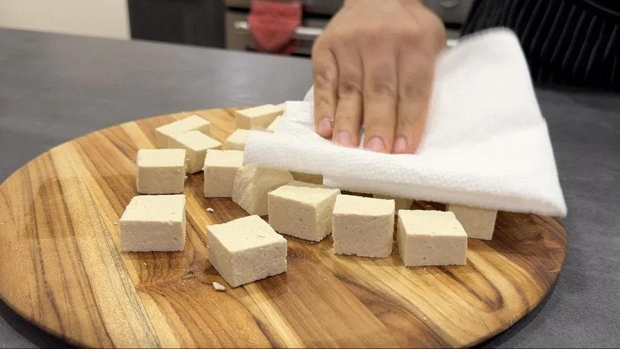 Hand patting tofu cubes dry with paper towel on wooden cutting board.