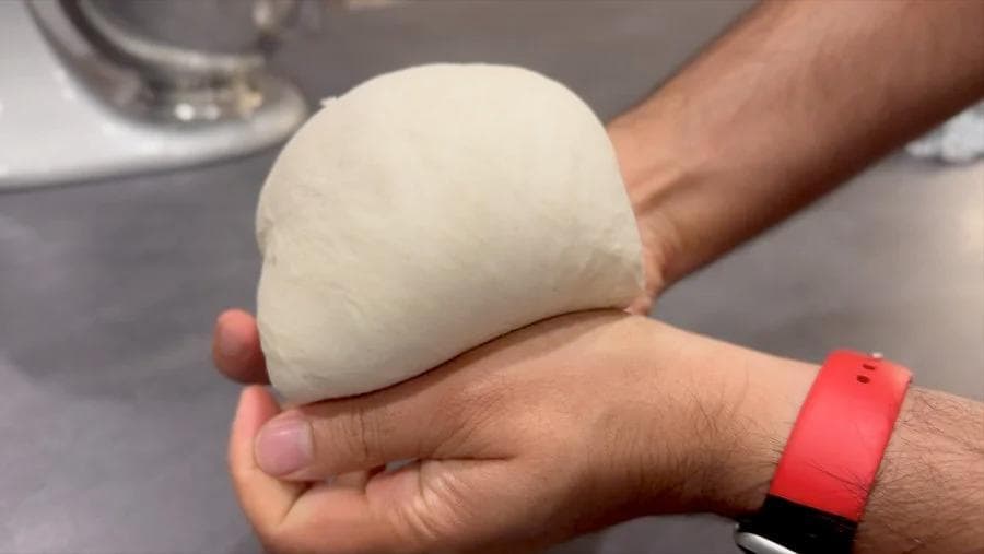 Hands holding a round ball of pizza dough with smooth, tight surface tension after kneading and folding.
