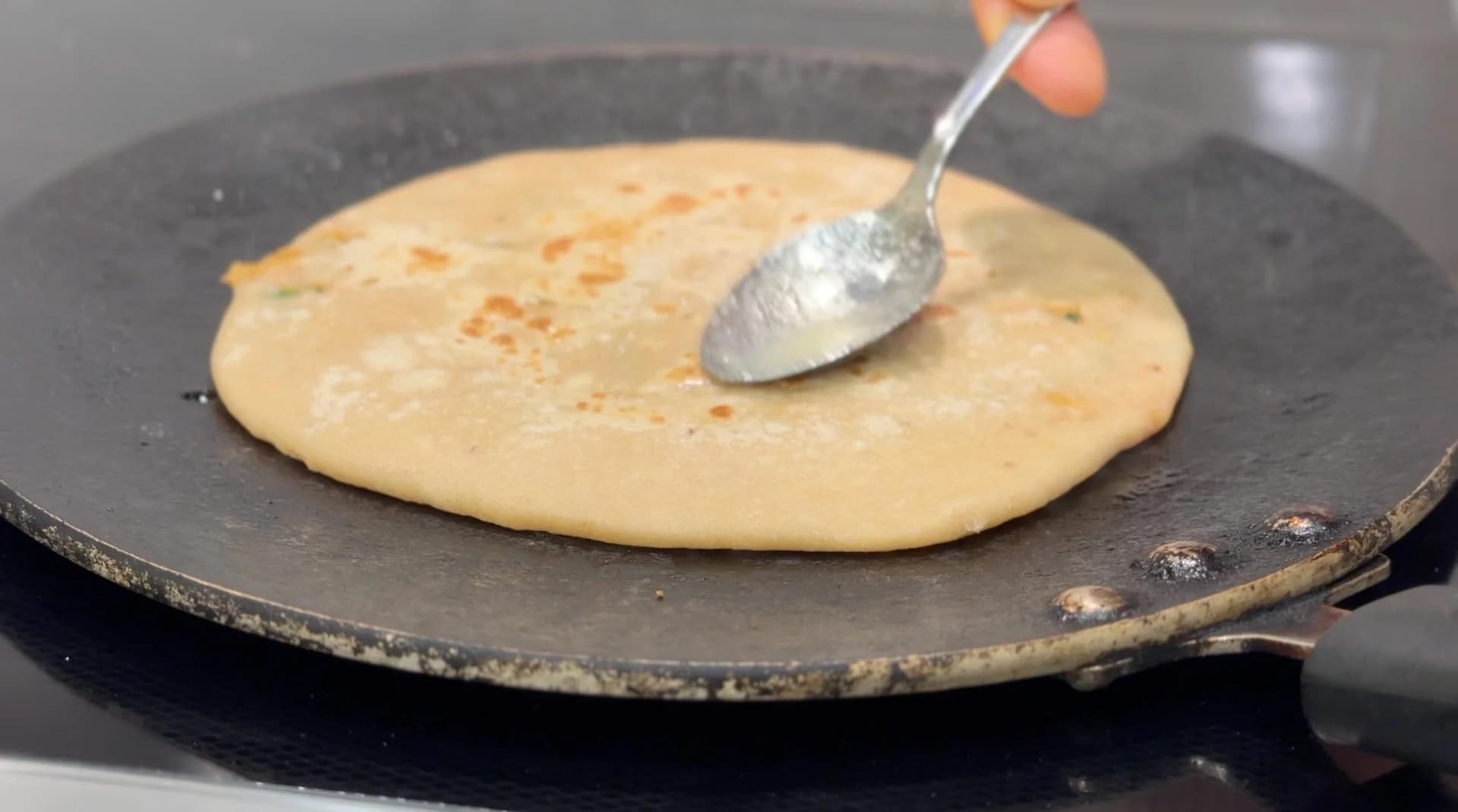 Gobi paratha cooking on a dark tawa with ghee being spread on top using a spoon.