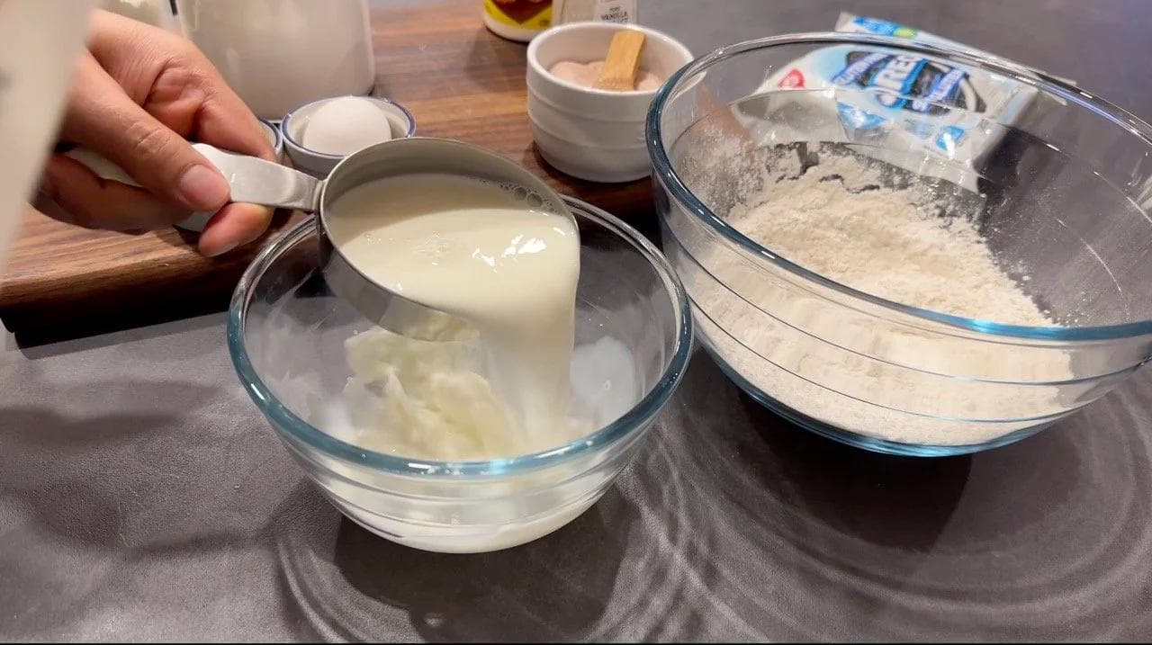 Wet ingredients being poured into a bowl with dry flour mixture, with crushed Oreos and measuring spoons visible.
