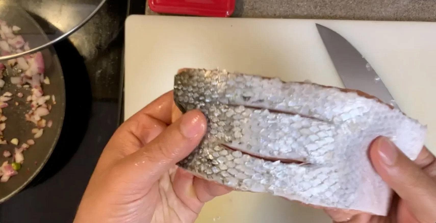 Hands descaling raw salmon fillet with a knife, showing the silvery scales being removed from the skin.