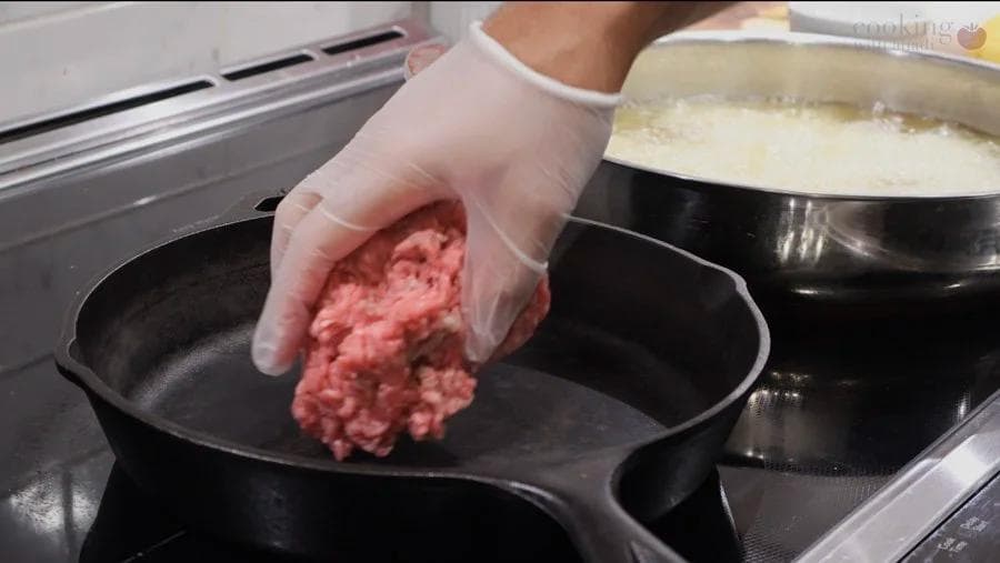 Hand in plastic glove adding ground meat mixture to a black skillet during preparation of perkedel daging.