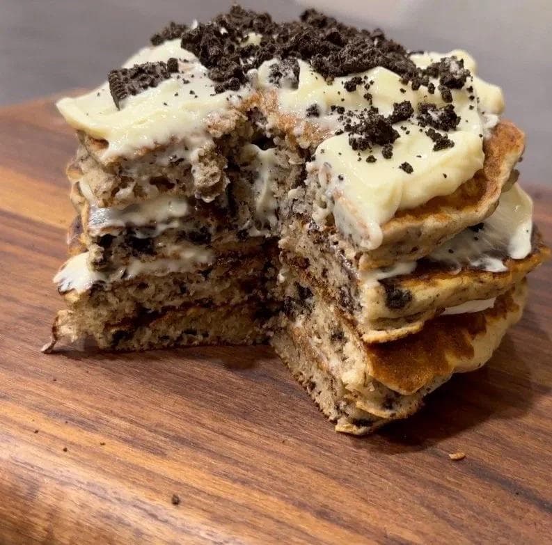 Stacked gluten-free Oreo cheesecake pancakes topped with cream cheese frosting and crushed Oreo cookies.