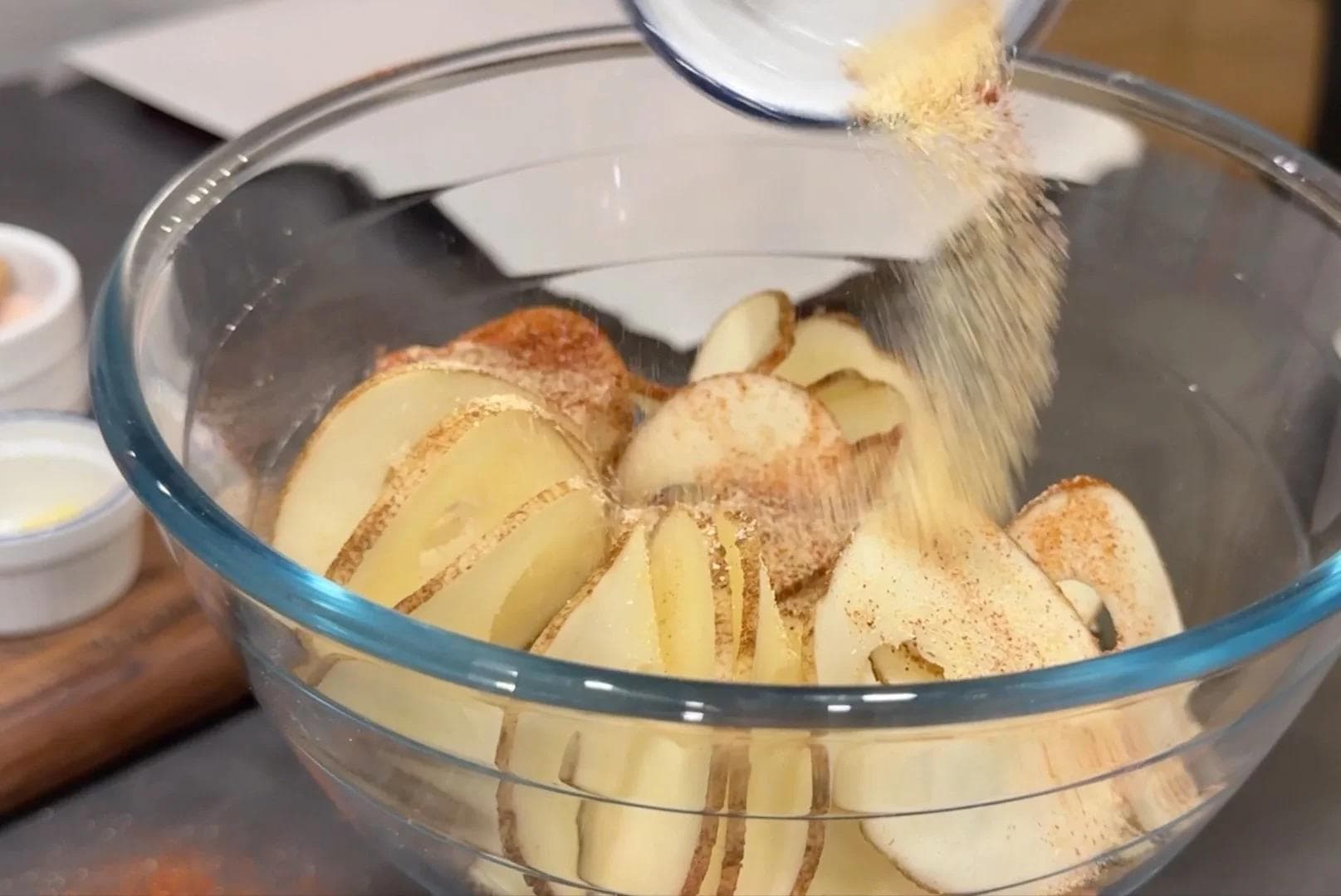Thinly sliced potatoes being seasoned with garlic powder, onion powder, and cayenne in a glass bowl.